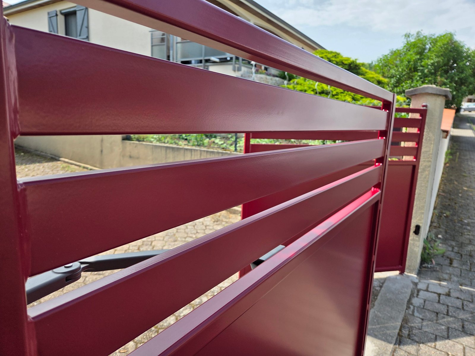 EDEL Électricien : Découvrez notre dernier projet de portail moderne en aluminium thermolaqué, ici présenté dans une élégante finition bordeaux. Alliant design épuré et robustesse, ce portail à lames horizontales ajourées offre un parfait équilibre entre intimité et esthétisme. Conçu pour s’intégrer harmonieusement dans un environnement résidentiel, il assure à la fois sécurité et style grâce à sa structure solide et son système d’ouverture motorisée discrètement intégré. Le rendu satiné du laquage reflète subtilement la lumière naturelle, ajoutant une touche de modernité à l’entrée de votre propriété. Un choix idéal pour ceux qui recherchent une solution à la fois durable, esthétique et contemporaine.